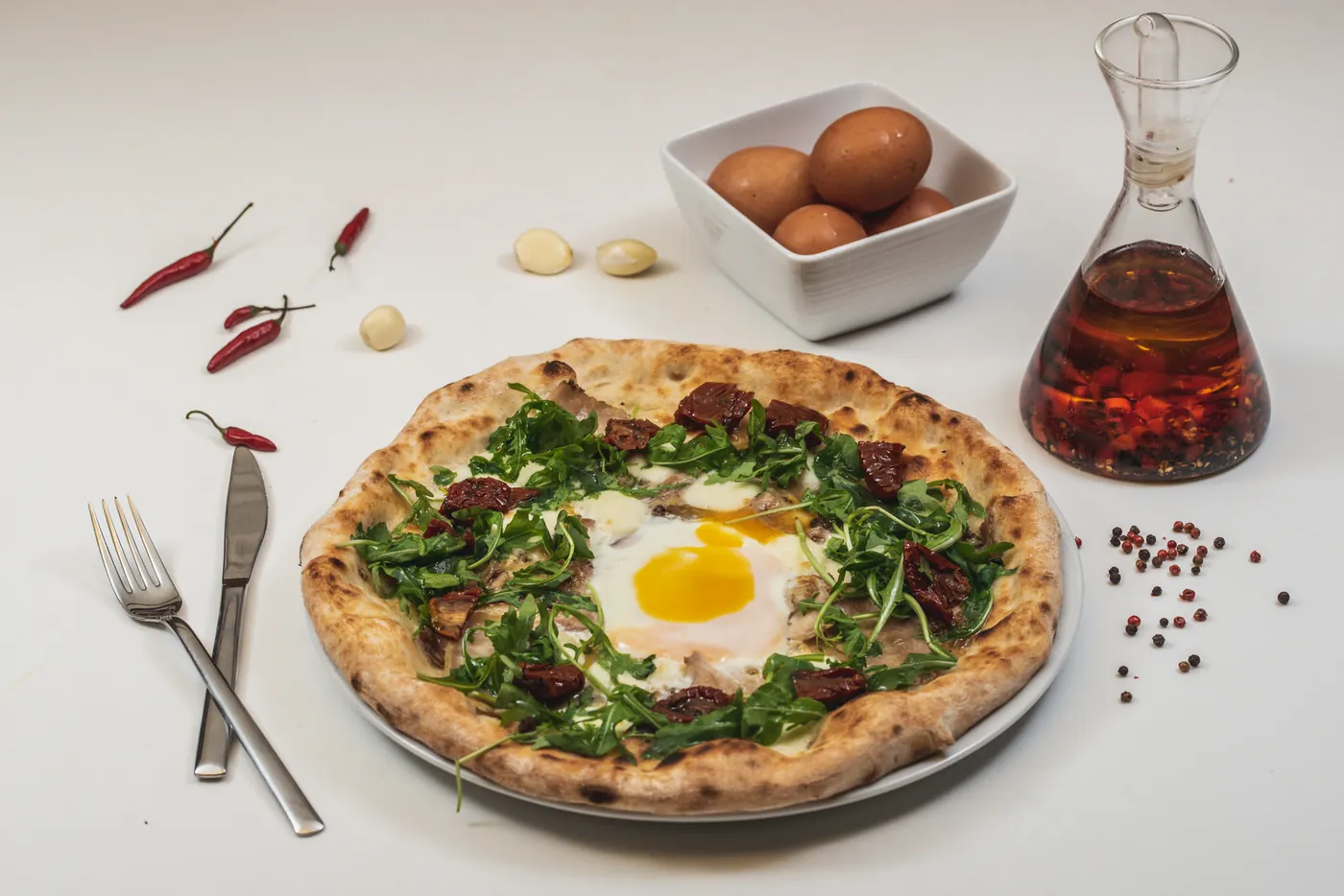 Pizza con un huevo en el centro, rúcula y tomate seco, entre guindillas, ajos y una aceitera