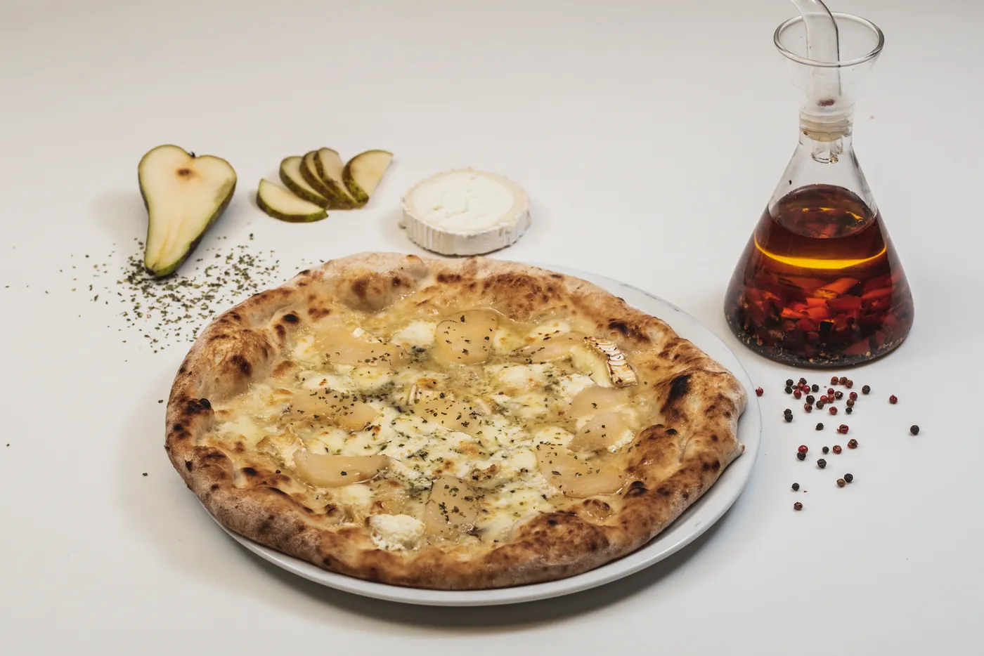 Pizza blanca con queso y pera, junto a media pera, rodajas de pepinillo y un queso de cabra