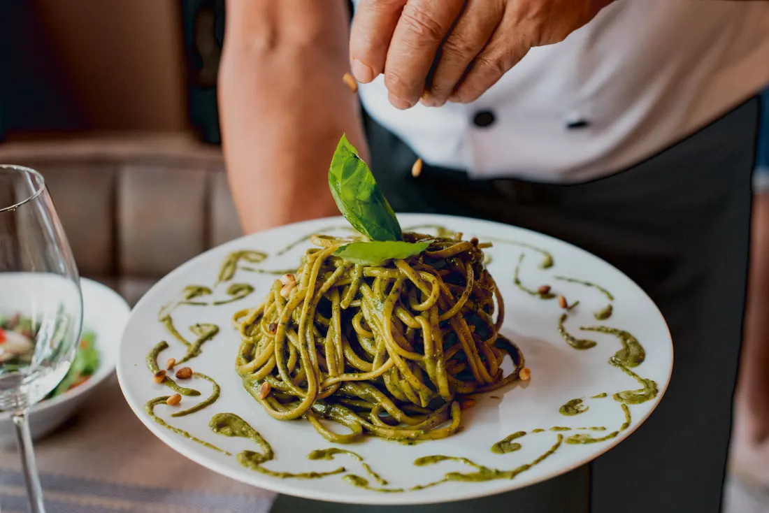 Una mano termina con piñones unos espaguetis al pesto coronados con una hoja de albahaca